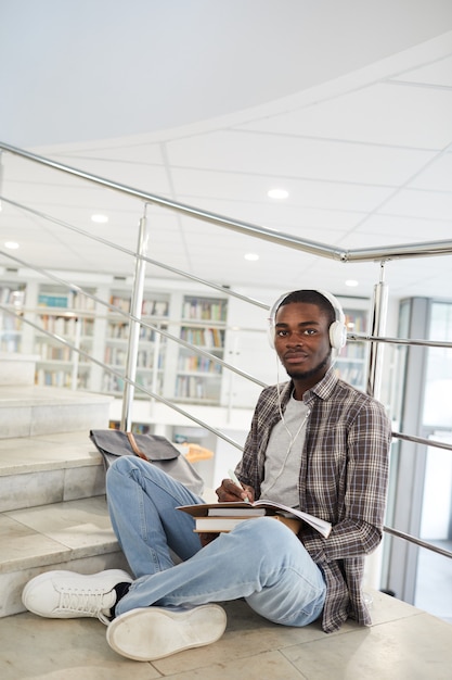 Vertical full length portrait of male African-American student listening to music while sitting cross legged on stairs in college and doing homework, 