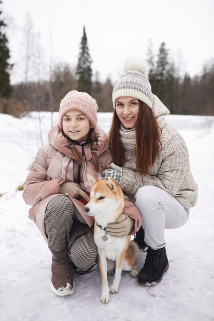 Vertical full length portrait of modern happy mother and daughter posing with pet dog and looking at camera while enjoying walk outdoors in winter forest