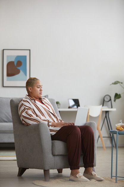 Vertical full length portrait of modern mixed-race woman using laptop while sitting in armchair at home, copy space