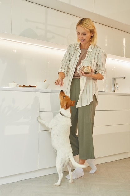 Вертикальный портрет в полный рост красивой женщины, угощающей собаку во время приготовления здорового завтрака в интерьере кухни