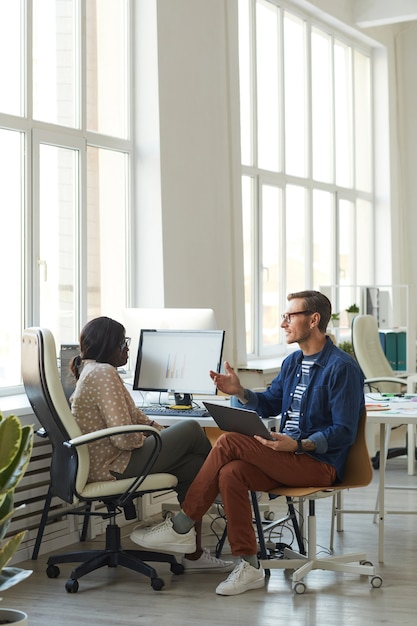 Vertical full length portrait of smiling contemporary businessman talking to African-American colleague while using computer in white office, copy space