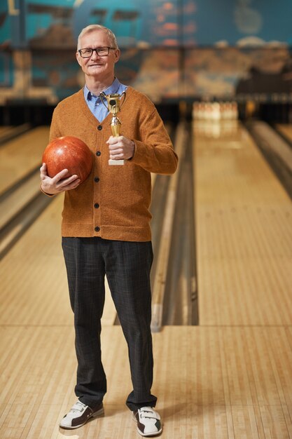 Ritratto a figura intera verticale di un uomo anziano sorridente che tiene in mano un trofeo e una palla da bowling mentre posa in una pista da bowling dopo aver vinto la partita, copia spazio