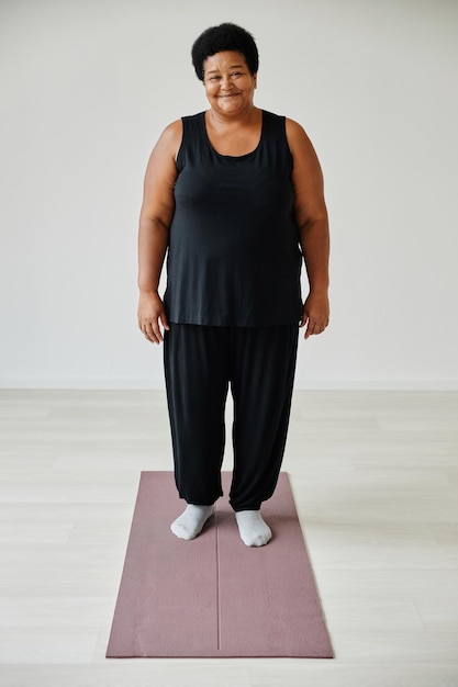 Vertical full length portrait of smiling senior woman standing on yoga mat while working out indoors