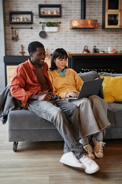 Вертикальный портрет молодой пары в полный рост с ноутбуком во время отдыха на диване в уютном домашнем интерьере