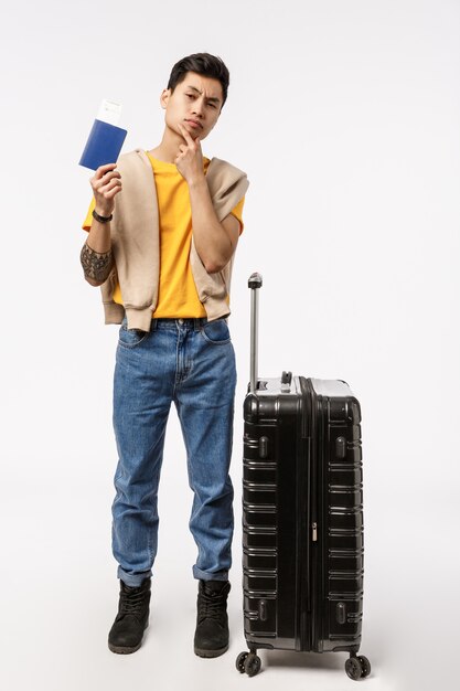 Vertical full-length shot thoughtful young asian guy standing near black suitcase, pack luggage decide travel abroad, study foreign country, touch chin thoughtful, holding passport, buy tickets