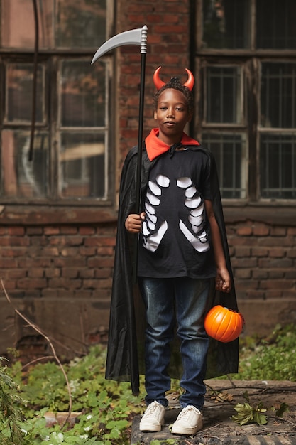 Vertical full shot portrait of cool African American boy dressed up as devil with red horns and scythe standing against old building 