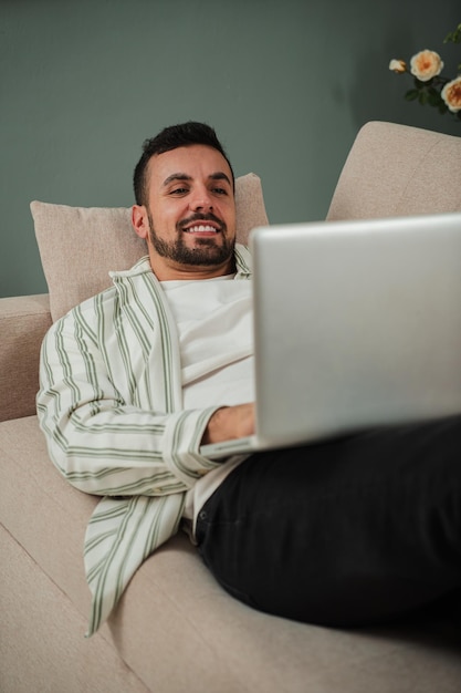 집에서 소파에 앉아서 노트북을 사용하는 수직적인 잘생긴 젊은 성인 남자 미소 짓는 남자