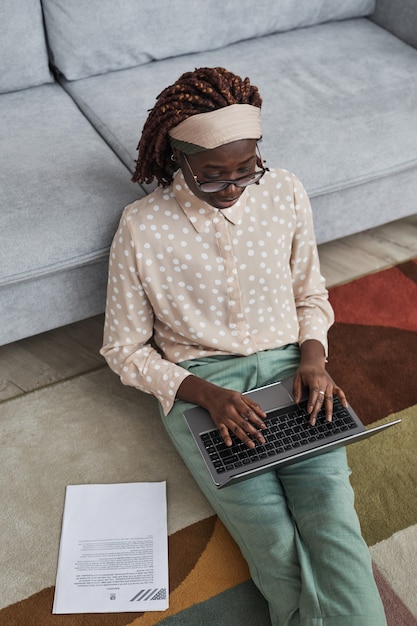 グラフィックカーペットの上に床に座って、ラップトップ、コピースペースを使用しながら自宅で仕事をしている現代のアフリカ系アメリカ人女性の垂直高角度の肖像画