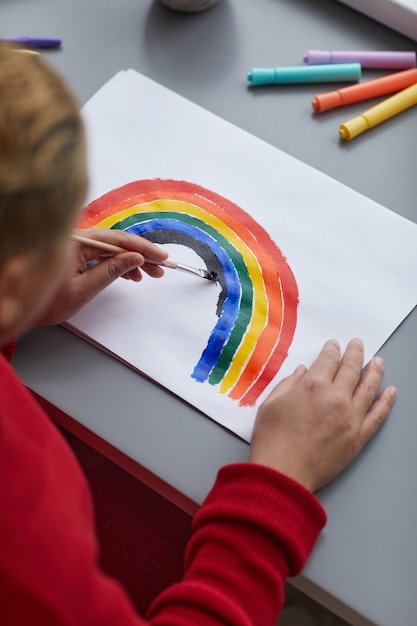 Vertical high angle view at modern mixed-race woman painting rainbow as LGBTQ symbol while sitting at desk at home, copy space