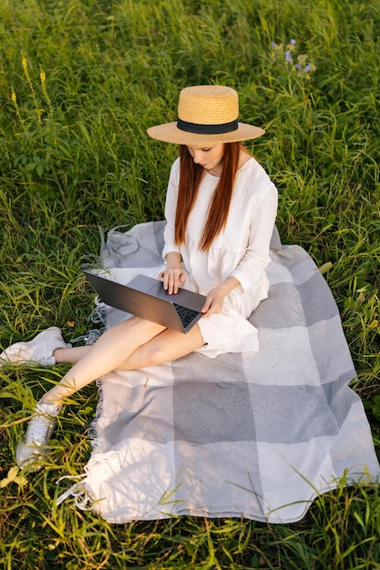 Vertical highangle view of focused female freelancer in straw hat and white dress working on laptop sitting on beautiful field of green grass