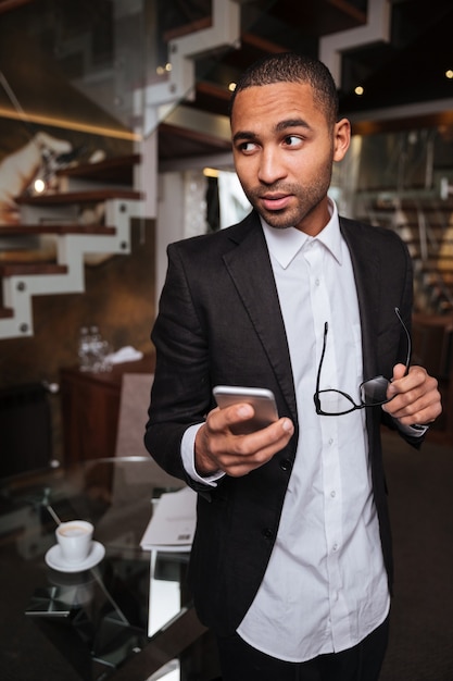 Vertical image of african man in suit with phone and glasses looking away in hotel