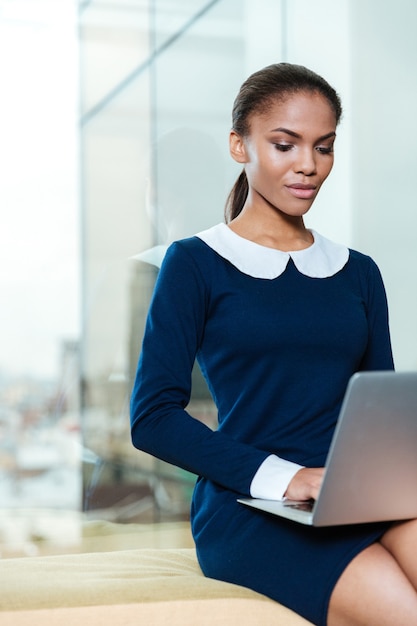 Vertical image of afro business woman in dress sitting near the window with laptop computer