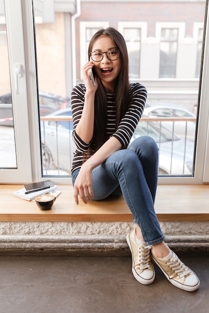 Vertical image of Asian woman talking phone on windowsill