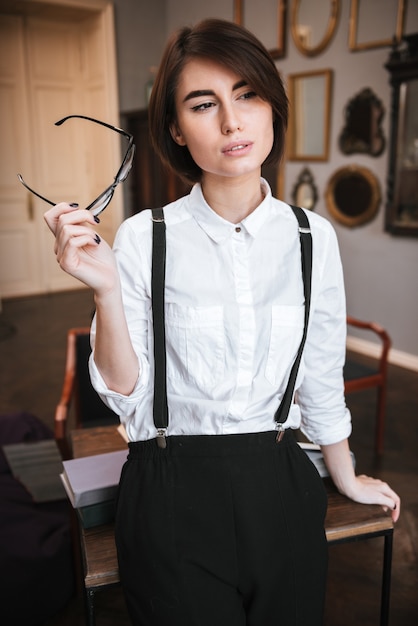 Photo vertical image of authoress in shirt standing near the table and holding eyeglasses in hand