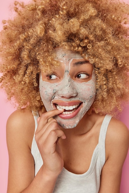 Vertical image of cheerful curious woman bites finger smiles positively and looks away applies beauty mask on face to remove bleakheads undergoes skin care procedures dressed casually poses indoor