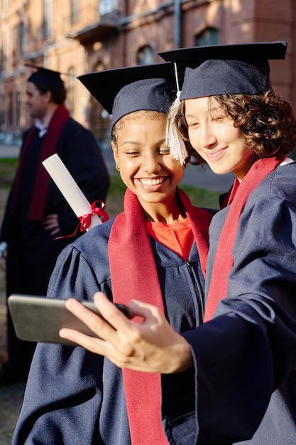 屋外でスマートフォンで自撮りポートレートを作る卒業式のガウンを着た女の子の垂直方向の画像