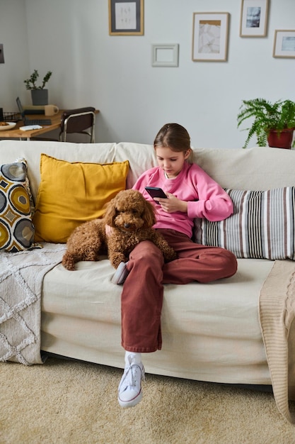 Vertical image of little girl sitting on sofa in the room and taking photo of her dog on smartphone