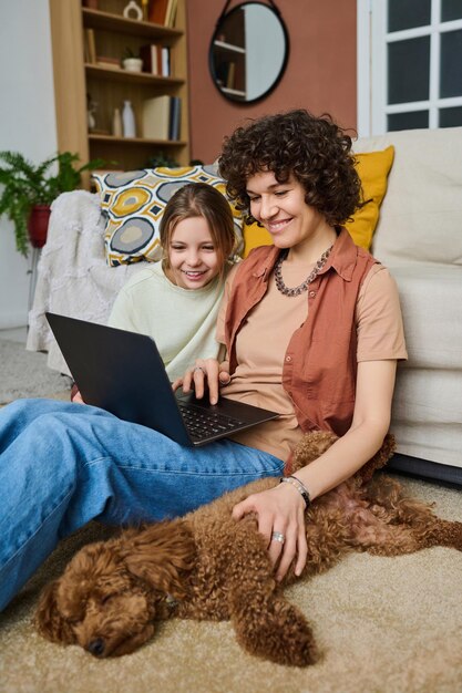 Vertical image of mom and daughter using laptop together with their dog resting near them on the flo