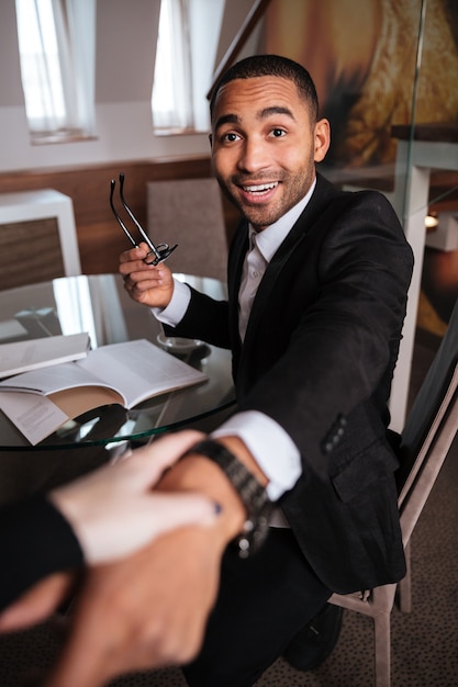 Vertical image of woman pulling happy african man in suit sitting by the table. First-person view