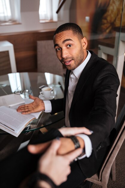 Vertical image of woman pulling young african man in suit sitting by the table. First-person view