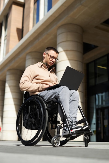 Вертикальное изображение молодого человека, сидящего в инвалидной коляске и использующего ноутбук в городе