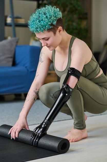 Vertical image of young woman with prosthetic arm preparing exercise mat for sport training at home