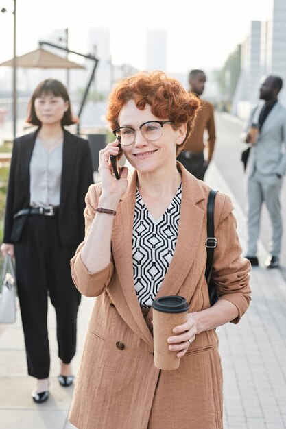 Vertical medium portrait shot of joyful mature woman with curly red hair walking along street talking on phone with someone after finishing workday in office