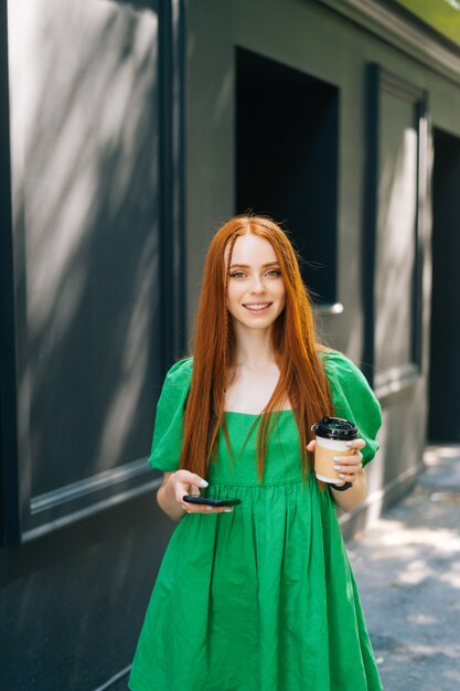 Vertical medium shot portrait of attractive young woman in green dress holding takeaway coffee cup u...