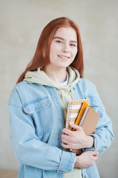 Vertical medium studio portrait shot of attractive young female\
student wearing casual outfit holding textbooks smiling at\
camera