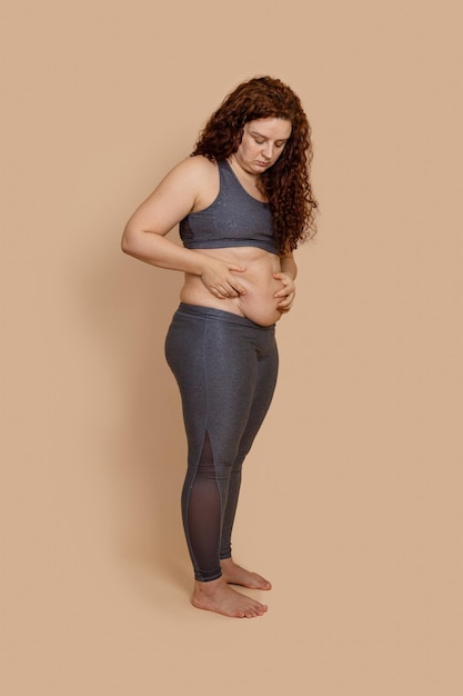 Vertical photo of barefoot depressed melancholic fat extra weight woman double chin looking on dangling belly Wearing gray sportswear Sad looking nonstandard Body positive Beige background