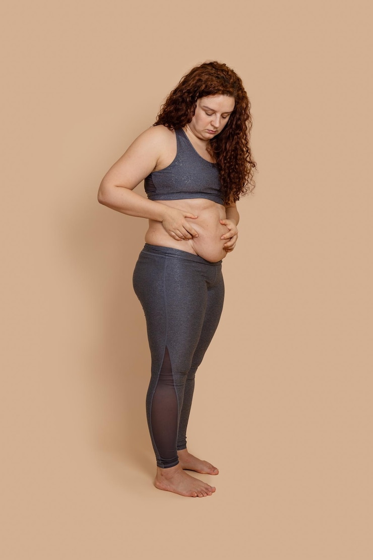Photo vertical photo of barefoot depressed melancholic fat extra weight woman double chin looking on dangling belly wearing gray sportswear sad looking nonstandard body positive beige background