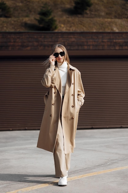 Vertical photo Beautiful carefree young teenage girl in gray raincoat and white jeans sneakers black glasses Walks around the outskirts of an Italian town Full length photo