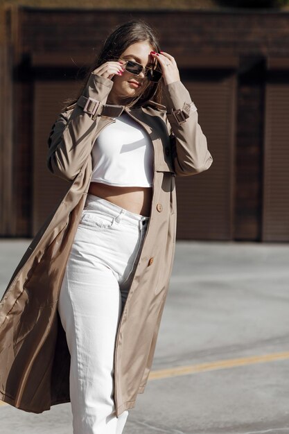 Vertical photo Beautiful carefree young teenage girl in gray raincoat and white jeans sneakers black glasses Walks around the outskirts of an Italian town