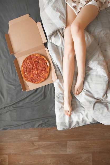 Vertical photo, box with pizza and girl's legs on the bed.