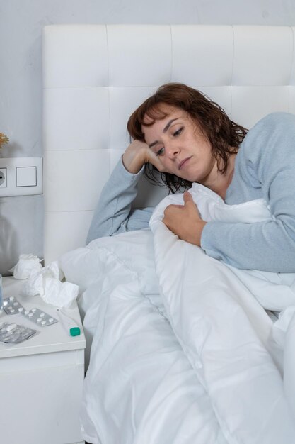 Vertical photo Caucasian mid adult woman wearing long blue pajamas sick in bed next to a bedside table with medication a thermometer and used tissues