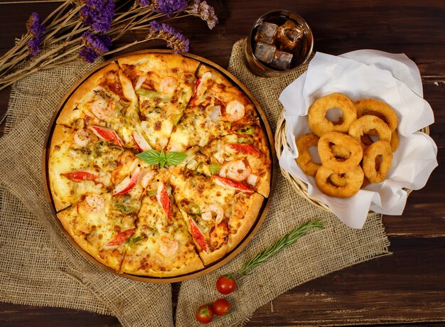 Vertical photo and close up on tasty homemade seafood pizza on sackcloth with dried flowers, cold beverage, Fried onions on wooden table. Food and Beverage Concept.