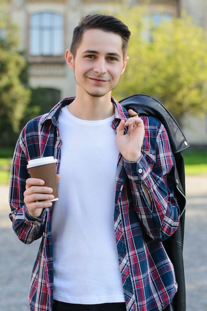 Foto verticale di un ragazzo calmo e allegro che tiene in mano una bevanda gustosa e una giacca di pelle nera sulla spalla