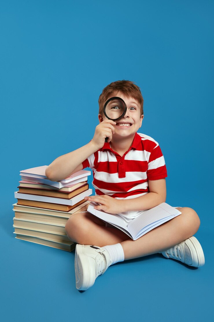 Photo vertical photo of excited smart boy looking at camera through magnifying glass and holding open book