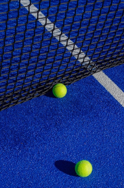 Vertical photo of two paddle tennis balls next to the net of a paddle tennis court.