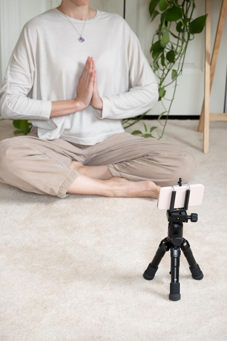 Vertical photo with selective focus on mobile with tripod, in the background a woman practicing yoga with copy space