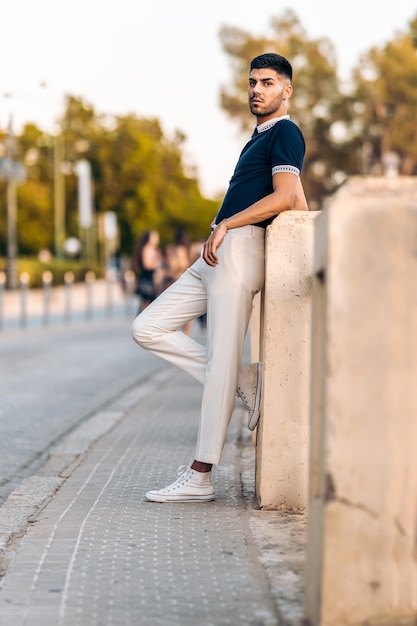 Vertical photo of a young caucasian man in modern clothes posing while leaning on a wall outdoors