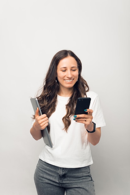 Vertical photo of a young woman is browsing on the phone while holding her laptop.