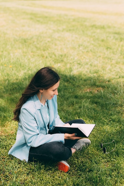 Vertical photo of a young woman sitting on grass and reading a book or her journal.