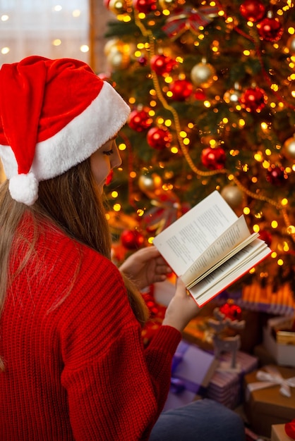 Foto foto verticale di una giovane donna seduta vicino all'albero di natale decorato e che legge un libro atmosfera calda e accogliente, magico momento felice a casa