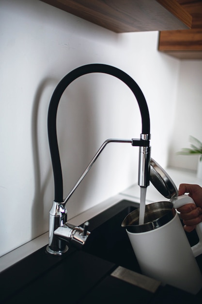 Фото Вертикальное изображение мужской руки, держащей электрический чайник с открытым верхом и наливающей немного воды из-под крана. необходимо нагреть воду. современная кухня с хорошей техникой и сантехникой