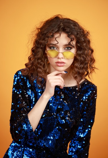 Vertical portrait of adorable curlyhaired girl put her finger to her chin on orange background High quality photo