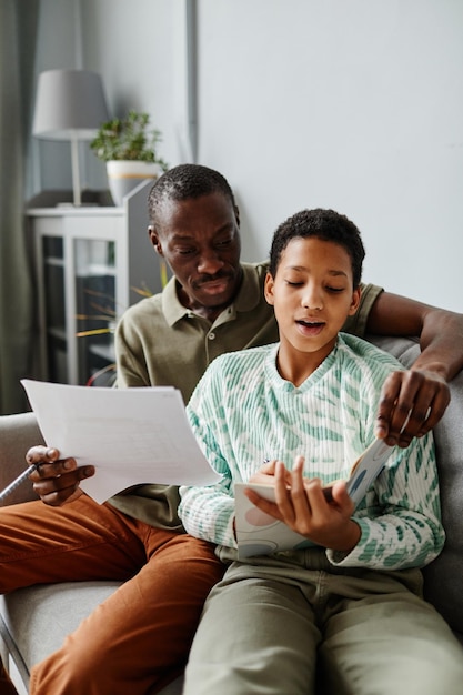 Vertical portrait of african american man helping daughter doing homework while sitting on sofa at home