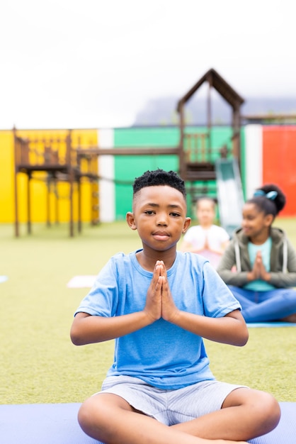 Vertical portrait of african american schoolboy practicing yoga meditation in schoolyard copy space