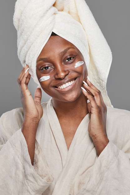 Vertical portrait of beautiful African-American woman enjoying skincare and looking at camera with face cream or moisturizer