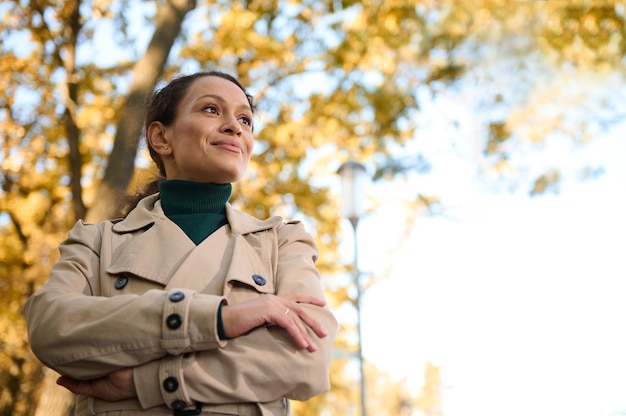 Vertical portrait of a beautiful confident business lady, attractive woman smiles looking away and stands with crossed arms on the autumnal nature background. Copy space